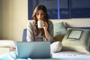 Woman sitting on bed sipping coffee looking at laptop