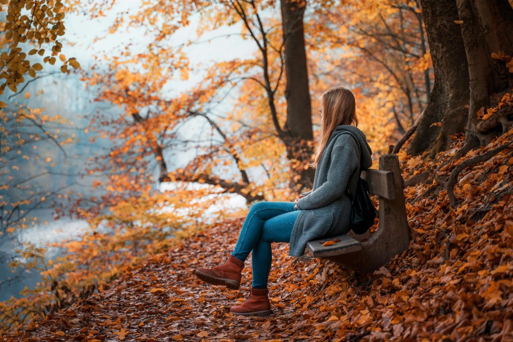 Woman sitting on bench beneath fall leaves looking at water.